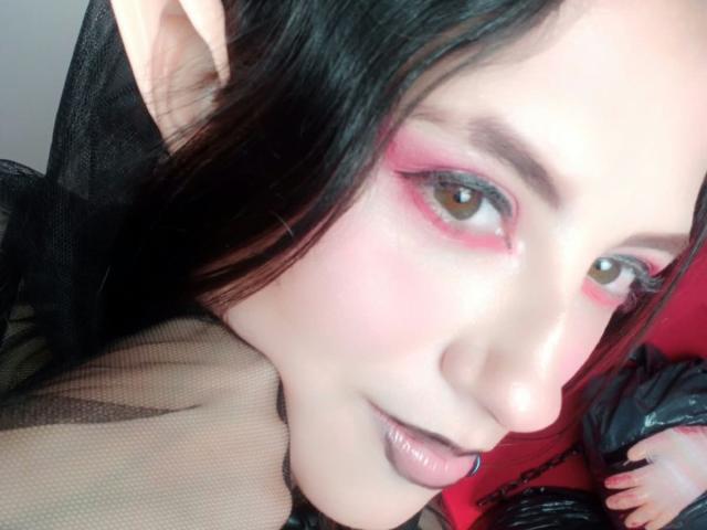 MaryCruzz's face with pale skin, pink eyeshadow, black lipstick, and pointed elf ears, lying down, wearing black mesh, red background, and black puffy jacket in the shot.