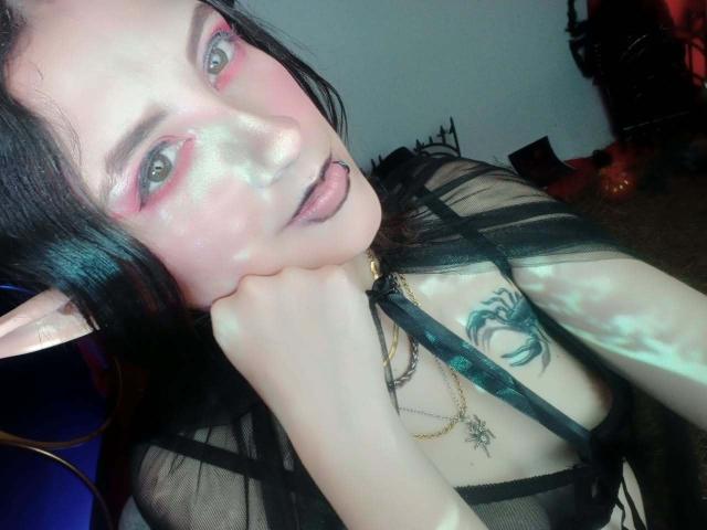 MaryCruzz with pale skin, pink eyeshadow, black lipstick, elf ears, black sheer top, hand on cheek, tattoo on chest, and gold necklace, looking at camera with tongue out.