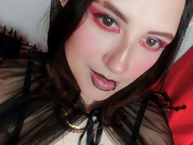 MaryCruzz with pink eyeshadow, black lipstick, tongue out, dark brown hair, sheer black top, gold necklace, pale skin, looking at camera, red cloth in background.
