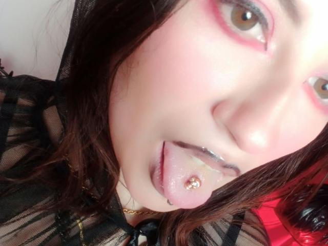 MaryCruzz's face with pink eye makeup, brown eyes, black mesh top, and tongue out, showing a gold ball-stud in her tongue, with saliva on her tongue and lips.
