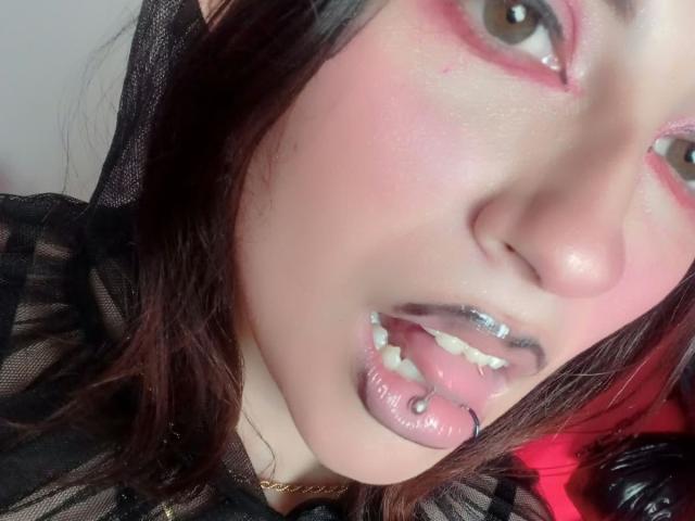 MaryCruzz's face with pink eye makeup, silver lip ring, and tongue piercing, wearing black lace, brown hair, and light skin, looking up with slightly parted lips.