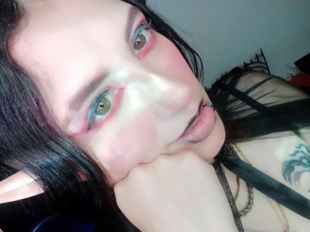 MaryCruzz with pale skin, black hair, green eyes, pink eyeshadow, black lipstick, tongue out, black strap, pointed elf ears, tattoo on right shoulder, hand under chin, shiny skin.