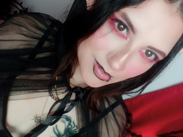 MaryCruzz with black sheer top, pink eye makeup, black lip ring, tongue out, green tattoo on chest, black hair, pale skin, and red background.