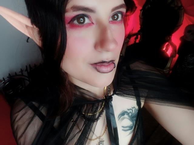 MaryCruzz with elf ears, pink eyeshadow, black lipstick, tongue out, black sheer top, gold chain, tattoo on chest, dark hair, red light in background.