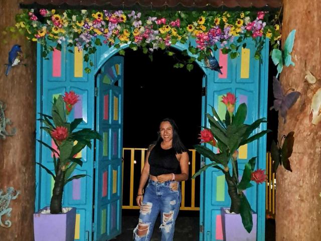 Jade Foster está de pie frente a unas puertas de color azul brillante, adornadas con flores, vistiendo una camiseta corta negra y unos vaqueros azules rotos, con una sonrisa, y de complexión media, cabello oscuro y piel morena..