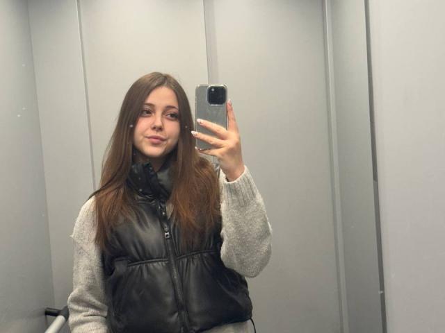 MaidaCain with long brown hair, wearing a gray sweater and black leather vest, taking a mirror selfie with a smartphone in a changing room.