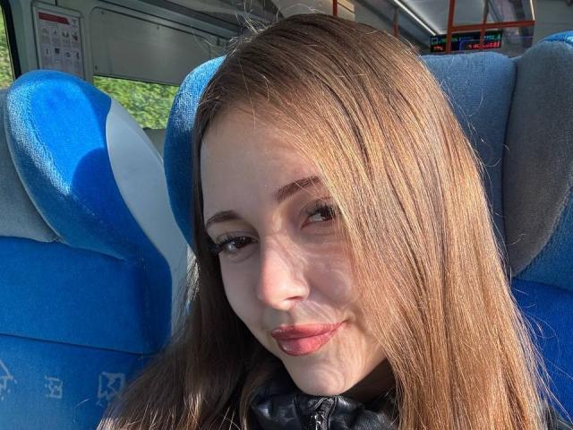 MaidaCain's face in a bus, blue and gray seat, long brown hair, light skin, subtle smile, wearing a black leather jacket, sunlight highlighting her features.