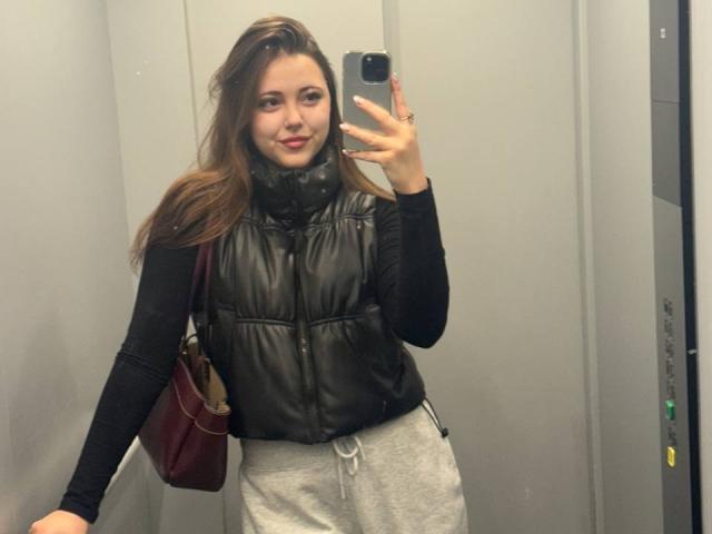 MaidaCain with brown hair, black puffy vest, black long-sleeve shirt, gray sweatpants, maroon bag, holding phone, smiling in mirror selfie, showing a cute face and ass.