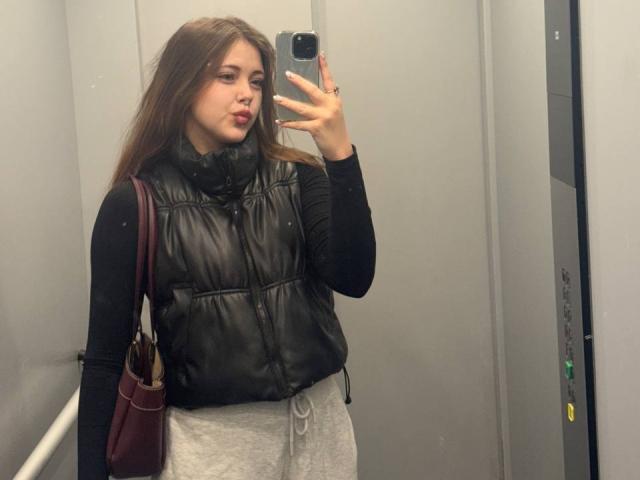 MaidaCain with long brown hair, black puffy vest, black long-sleeve shirt, grey sweatpants, and maroon bag, taking a mirror selfie in an elevator, showing a pouty face.