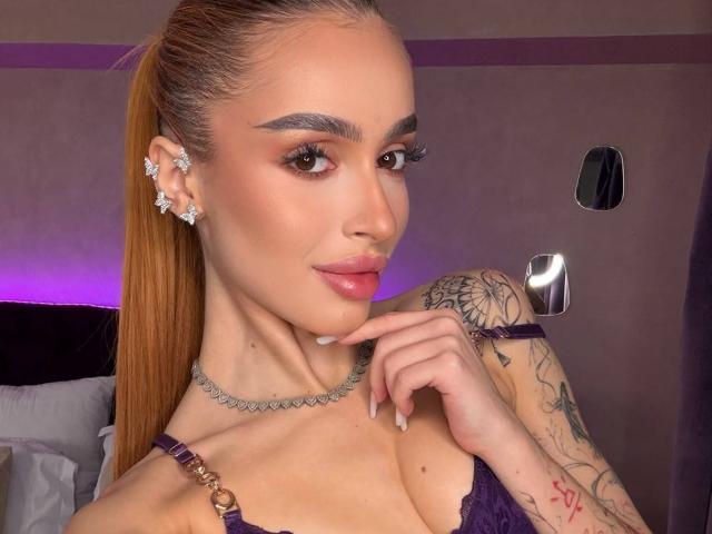 MarissaFex com rabo de cavalo castanho, sobrancelhas escuras, lábios rosados, brincos de diamante, lingerie roxa, braço tatuado, dedo no queixo, colar de prata, delineador rosa e iluminação roxa. Seios..