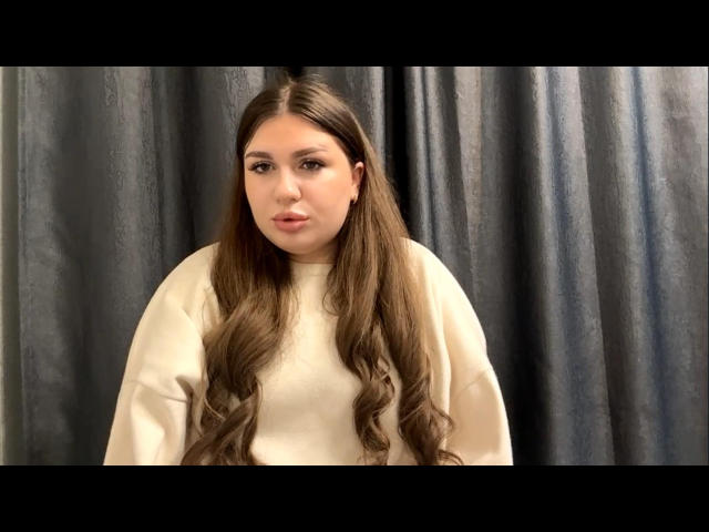 NormaLampey with long brown wavy hair, white sweater, and subtle makeup, standing against a dark curtain, giving a neutral expression. Boobs are not visible.