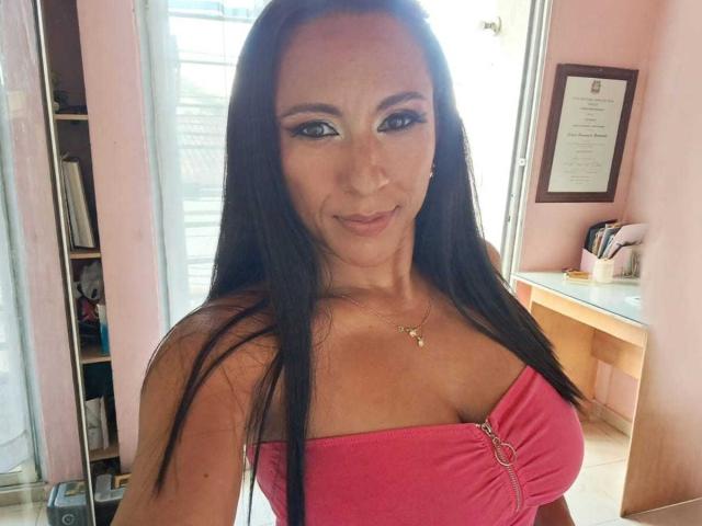 Jade Foster, con cabello negro largo, ojos azules, un top rosa sin tirantes que muestra sus grandes senos, una ligera sonrisa y un collar dorado, de pie en una habitación iluminada por el sol..