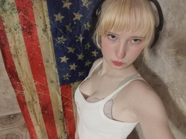JennifferBreaz with blonde bob, wearing white tank top, showing small tits in gray bra, looking up, against American flag, wearing black headphones, pale skin, pouty lips.