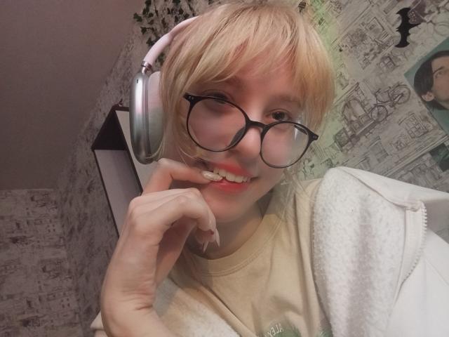 JennifferBreaz with blonde hair, glasses, and pink headphones, smiling with finger on chin, wearing a beige shirt and white hoodie, showing her teeth.