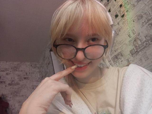 JennifferBreaz with blonde hair, glasses, and headphones, biting her finger with a cute smile, wearing a beige shirt, in a selfie. Her nose is slightly red.