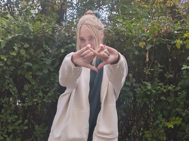 JennifferBreaz with blonde hair in a bun, wearing a white jacket, making a heart shape with her fingers, green leafy bush behind her, blue shirt visible underneath.
