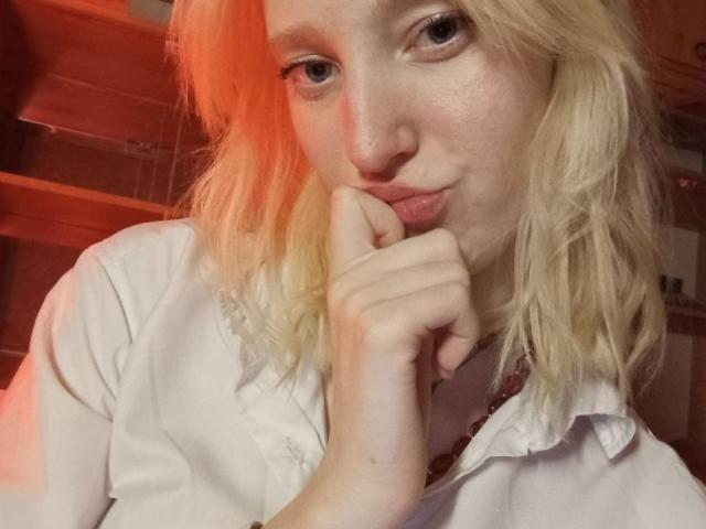 JennifferBreaz with pale skin, blonde wavy hair, pouty lips, and a white shirt, biting her finger, looking at the camera, with a red light on the left.