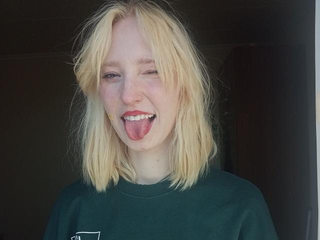 JennifferBreaz has blonde, messy hair, is winking with her tongue out, and wearing a green shirt, showing her pink tongue and white teeth.