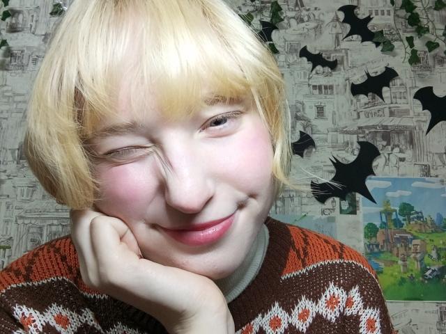 JennifferBreaz with blonde hair, winking, smiling, pink cheeks, hand under chin, wearing brown and white patterned sweater, background with bat cutouts and city illustrations.