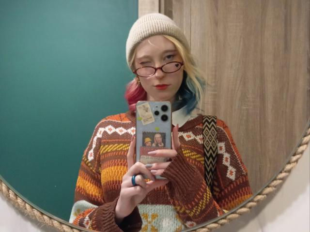 JennifferBreaz with blonde and blue hair, wearing glasses, a beige beanie, and an orange patterned sweater, taking a mirror selfie with a phone. Boobs, ass not visible.