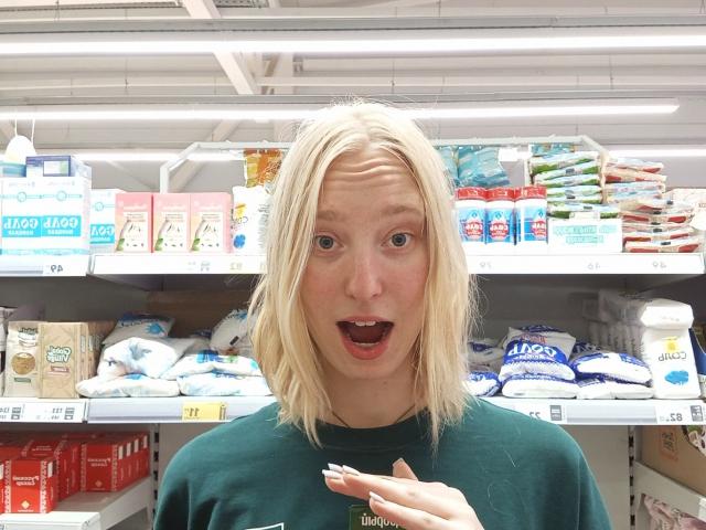 JennifferBreaz with blonde hair, green shirt, shocked expression, mouth open, eyebrows raised, hand up, in supermarket aisle, surrounded by packaged food, bright fluorescent ceiling lights.