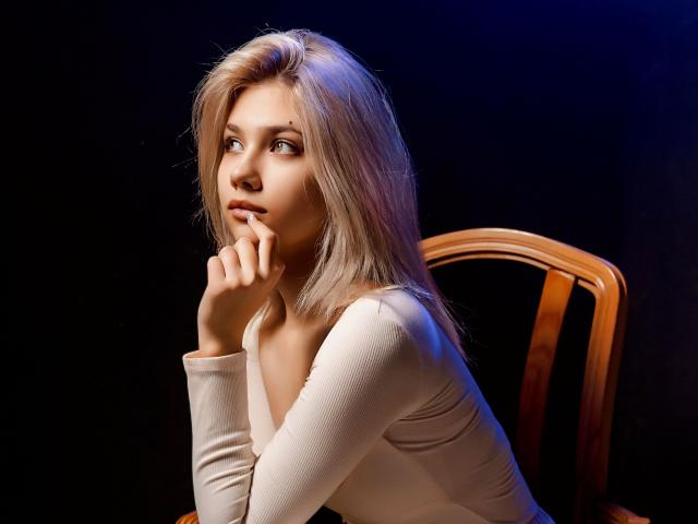 DataSley with blonde hair, thinking, has her hand near her mouth, wearing a tight beige top, sitting on a wooden chair, dark background, blue light on her right.