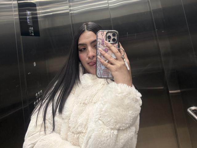 LunnaBlossom, con lunghi capelli neri, indossa un cappotto bianco soffice e sta scattando un selfie in un ascensore di metallo, tenendo un iPhone con una custodia rosa a motivi. Ha un'espressione accigliata..