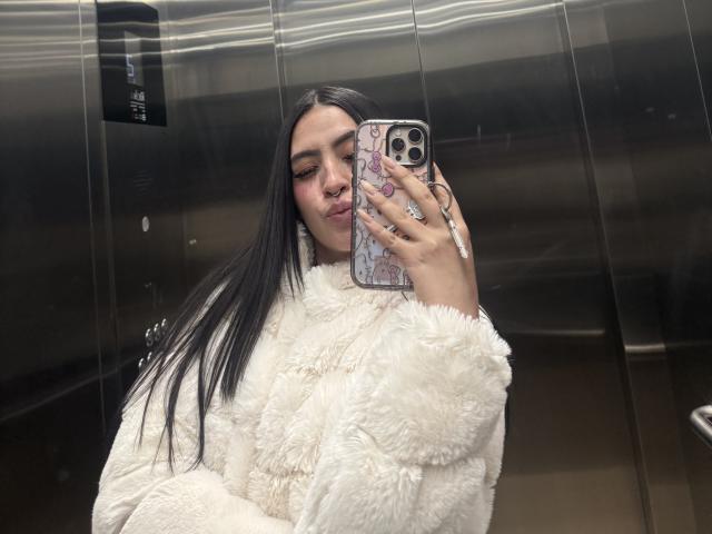LunnaBlossom with long black hair, wearing a fluffy white coat, taking a mirror selfie in an elevator, holding a phone with a pink and purple case.