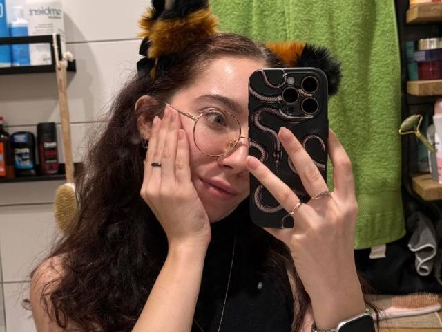 ClaireSpisak with wavy brown hair, glasses, and furry cat ears, holding a phone with a snake pattern case, covering her mouth, wearing a black turtleneck.