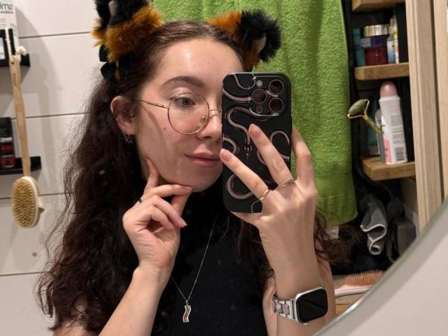 ClaireSpisak with cat ears, wearing glasses, holding a phone, black turtleneck, ring on finger, smartwatch, green towel, curly brown hair, slightly smug expression, taking a mirror selfie.