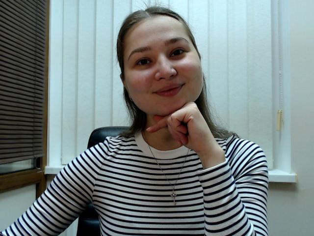 SharlotWings with brown hair, wearing a striped shirt, resting her chin on her hand, smiling slightly. Her pussy is not visible. She's in an office with blinds.