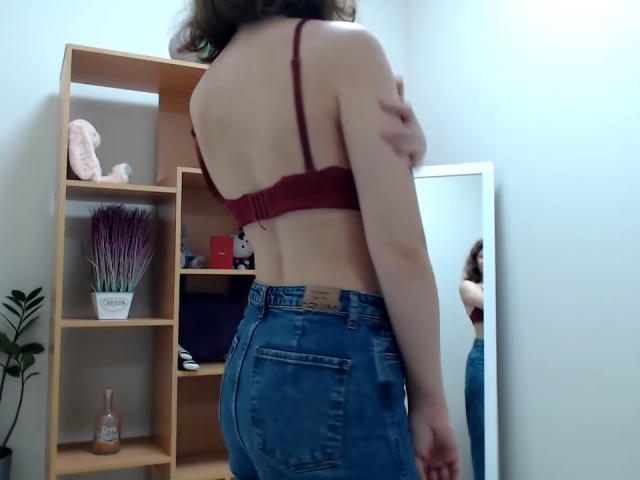 Steisii with her back to the camera, wearing a red bra and blue jeans, standing in front of a mirror, her hand on her shoulder, showing her ass.