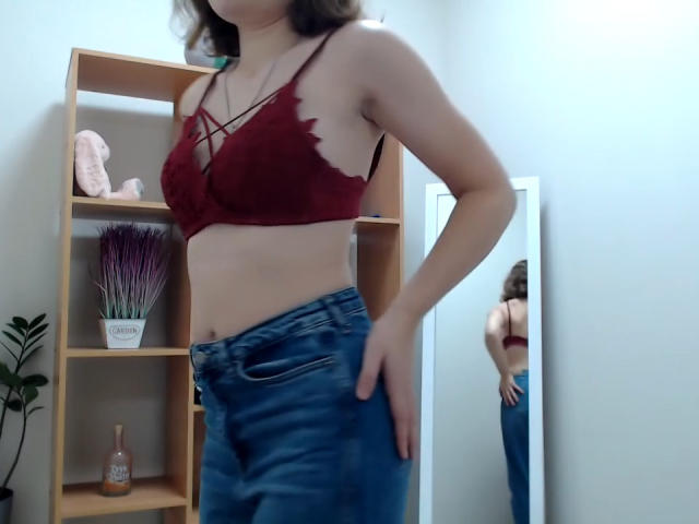 Steisii in a red lace bralette, showing her tits, with blue jeans, standing sideways, reflected in a mirror, hand on hip, wavy brown hair, white wall background.