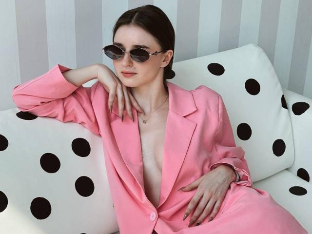 TeofilaMaupins with pink blazer, open chest, small tits, sunglasses, black polka dot couch, hand on chin, necklace, casual pose, light skin, brown hair, relaxed expression.