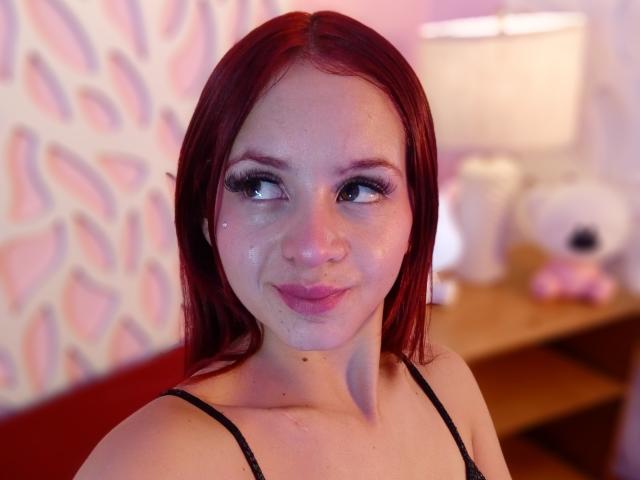 LesleyGray with red hair, wearing a black spaghetti strap top, has a shiny face with visible semen on her chin and nose. She has long eyelashes and a small smile.
