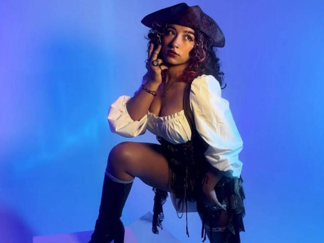 KanelaMiller with dark skin, curly red hair, pirate hat, white puffy shirt, black corset, knee socks, squatting, holding phone, blue light, looking up, showing big tits.
