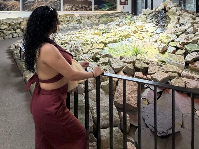 Aurora Ferretti, avec ses cheveux noirs bouclés, sa robe rouge sans haut, ses formes généreuses, et dos à la caméra, se tient près d'une fontaine ornée de rochers, tenant une rambarde en métal noir..