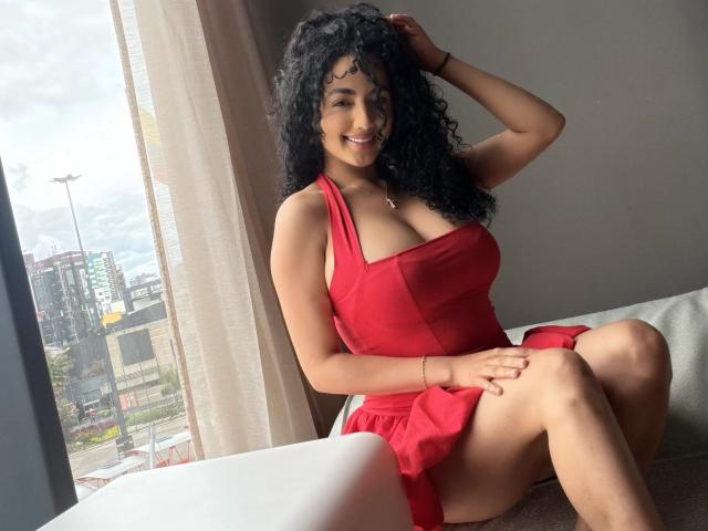 AuroraFerretti with curly black hair, wearing a red dress, sitting on a couch, smiling, hand in hair, showing her big tits, red garter, and legs spread.