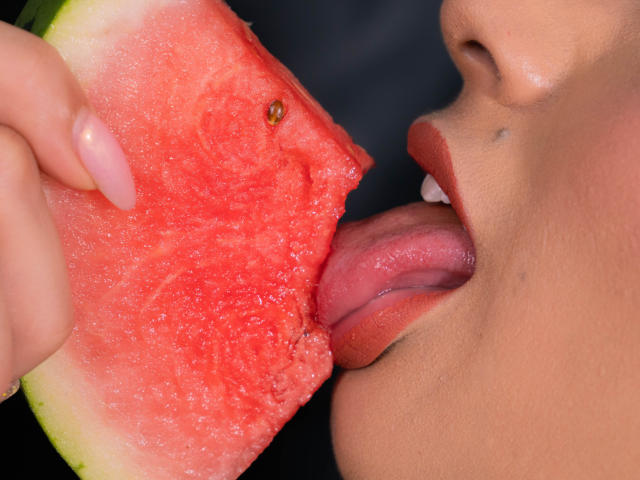 JasminKooper's tong hangt uit, terwijl ze een sappig stuk watermeloen likt, vastgehouden door haar roze gelakte vingers. Haar lippen zijn open, waardoor haar witte tanden zichtbaar zijn. Haar huid is bruin..