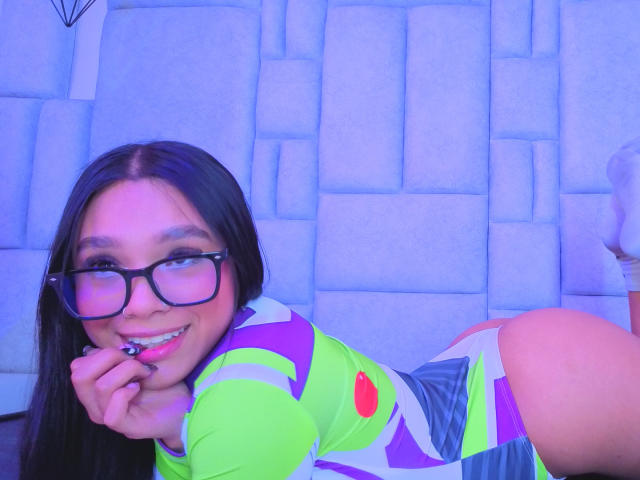 MiiaBoss with glasses, lying on her side, smiling, wearing a neon green and purple outfit, showing her butt, holding a lollipop, blue light background.