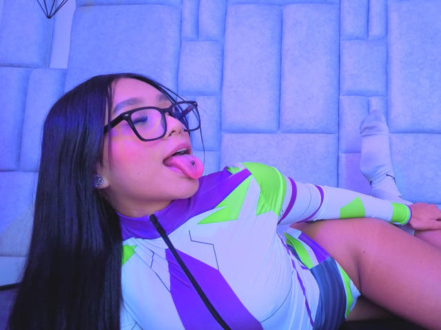 MiiaBoss with long black hair, glasses, and pink tongue out, wearing a purple and green futuristic outfit, lying back with legs up, showing her ass. Blue light.