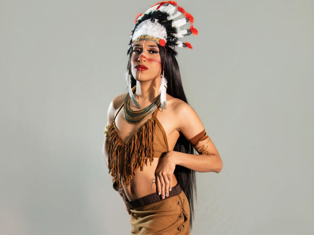 PaulaHills with long black hair, wearing a Native American-style headdress, brown fringed top, and skirt, showing her small tits and brown arm bands, standing confidently.