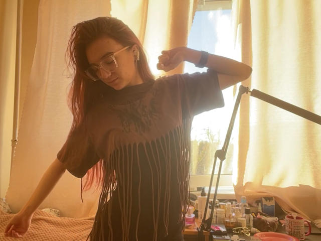 ThelmaSeilhamer with red hair, glasses, and striped shirt, standing in sunlit room, raising arm, wearing earbuds, desk with lamp and stuff in background, looking down.