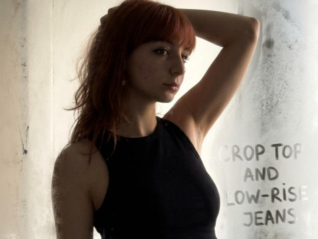 LeomaHaq with red hair, black sleeveless top, arm raised, looking to the side, soft light, slight shadow, freckles, subtle makeup, casual, confident, attractive, small tits.
