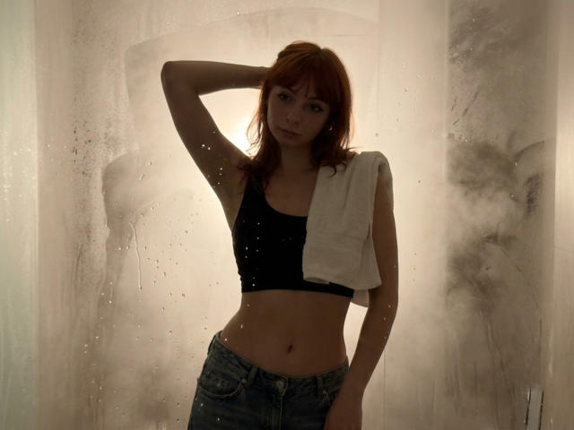 LeomaHaq with red hair, wearing a black sports bra, showing her small tits, and a white towel over her shoulder, standing in a steamy, wet bathroom, wearing jeans.