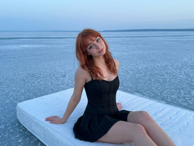 LeomaHaq with red hair, black strapless dress, sitting on white mattress on salt flat, hands on mattress, legs crossed, butt slightly raised, looking at camera, pale skin, calm expression.