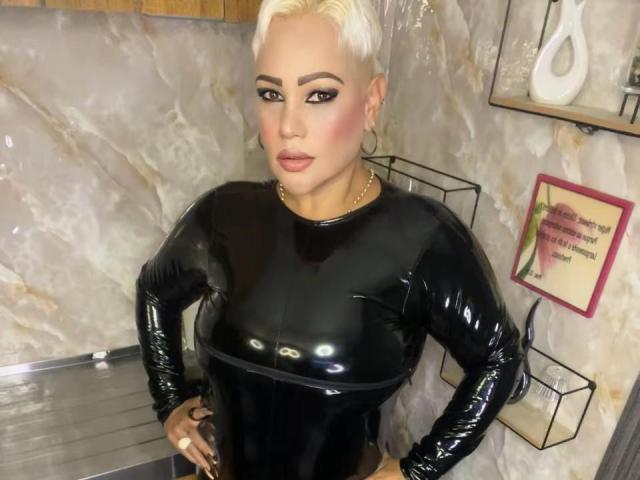 GianellaCox with platinum blonde hair, wearing a shiny black latex long-sleeve top, hands on hips, looking at camera, hoop earrings, makeup, marble wall background, confident expression.