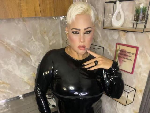 GianellaCox with platinum blonde hair, wearing a shiny black latex long-sleeve top, sucking her finger, large hoop earrings, and a black ring, looking seductively at the camera.