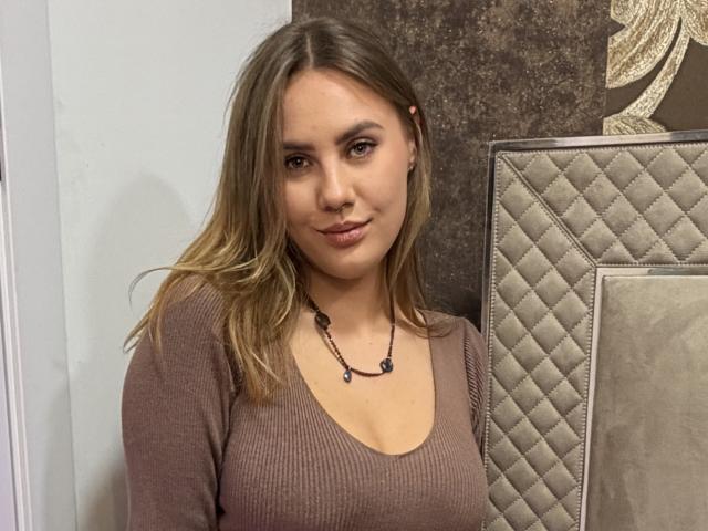 BettyLuv with blonde hair, brown eyes, and a nose ring, wearing a ribbed brown top and black necklace, smiling softly. Her boobs are medium and she's looking cute.