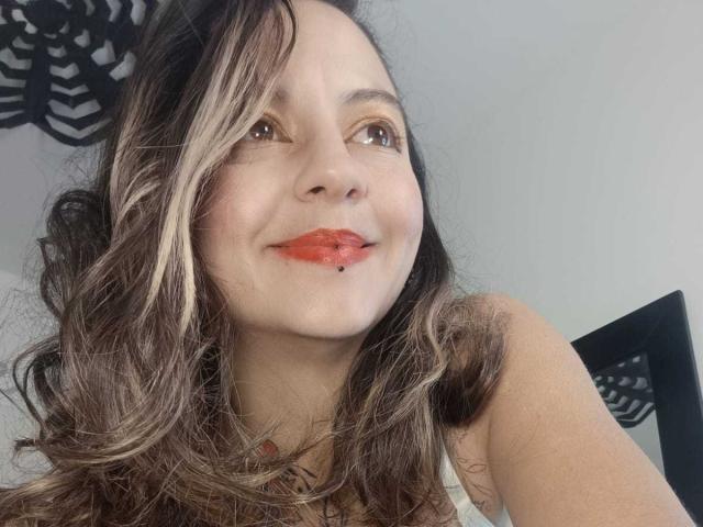 ArianaBlonde has wavy brown hair with blonde highlights, red lipstick, and a small beauty mark under her right eye, smiling at the camera. Her brown eyes are looking upward.